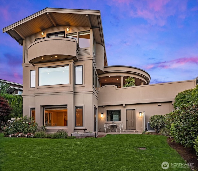 back of house at dusk featuring stucco siding, a lawn, and a balcony