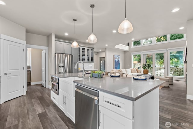 kitchen featuring a kitchen island with sink, a sink, appliances with stainless steel finishes, dark wood finished floors, and glass insert cabinets