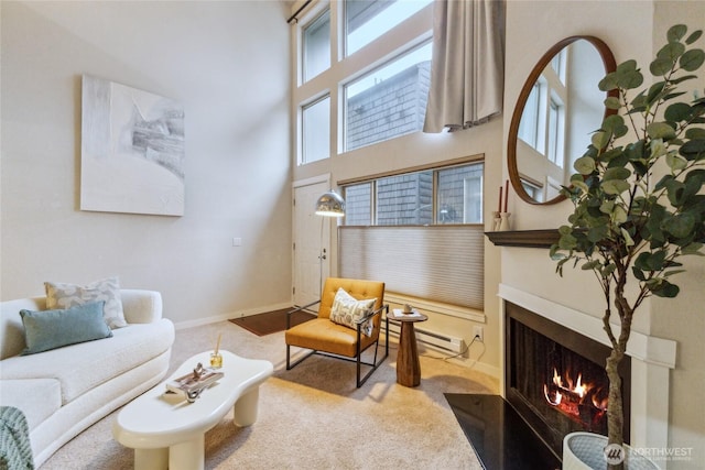 carpeted living room featuring a baseboard heating unit, baseboards, a high ceiling, and a lit fireplace