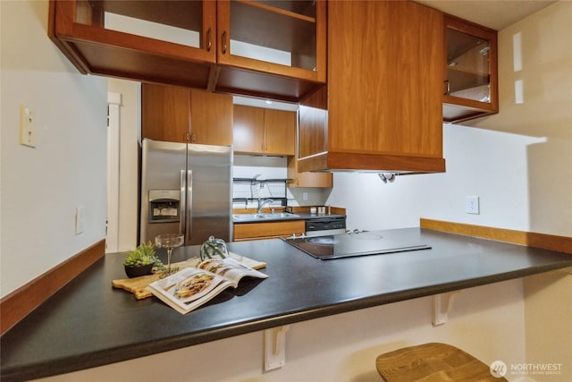 kitchen featuring brown cabinetry, a sink, glass insert cabinets, dark countertops, and stainless steel fridge