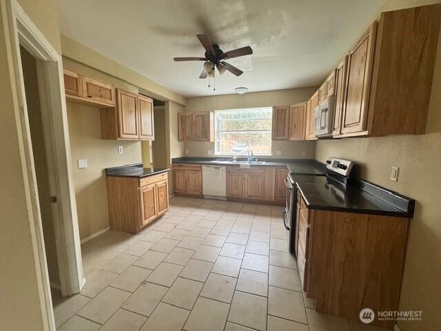 kitchen featuring brown cabinetry, a ceiling fan, dark countertops, appliances with stainless steel finishes, and a sink