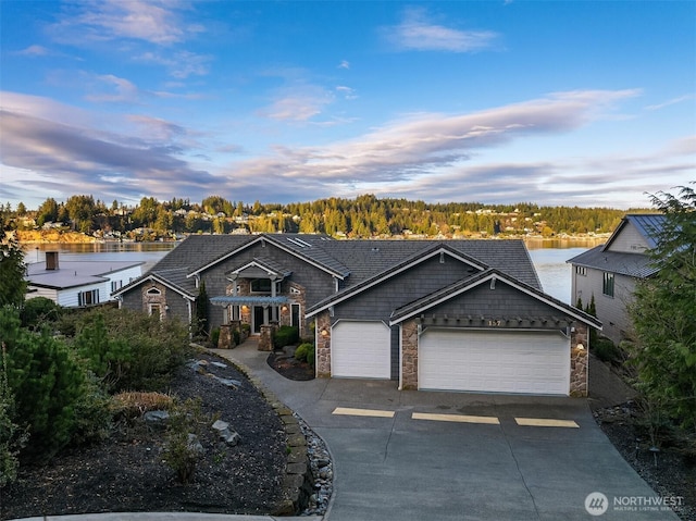 view of front of home with driveway, a garage, stone siding, a forest view, and a water view