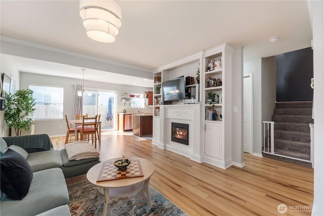 living area with crown molding, light wood finished floors, a glass covered fireplace, baseboards, and stairs