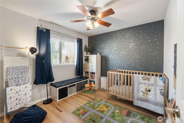 bedroom with an accent wall, wood finished floors, a nursery area, baseboards, and wallpapered walls