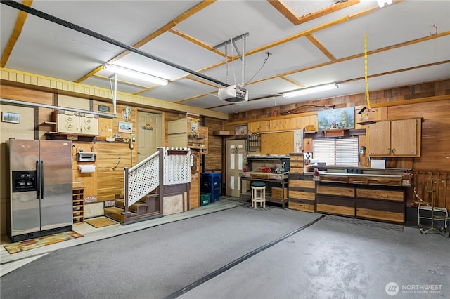 garage featuring a workshop area, a garage door opener, stainless steel fridge with ice dispenser, and wood walls