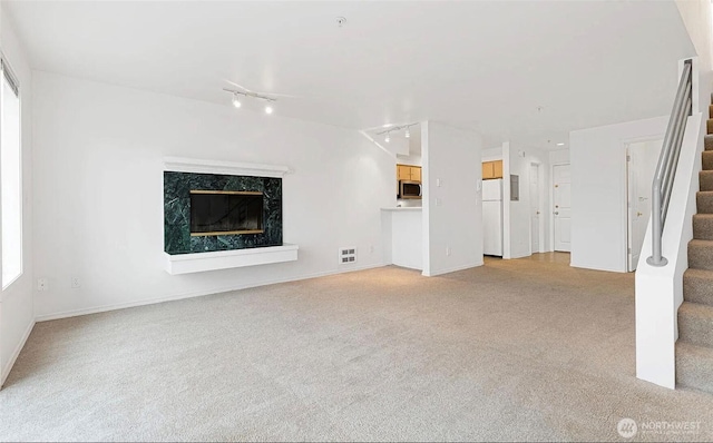 unfurnished living room featuring light colored carpet, a premium fireplace, visible vents, stairway, and rail lighting