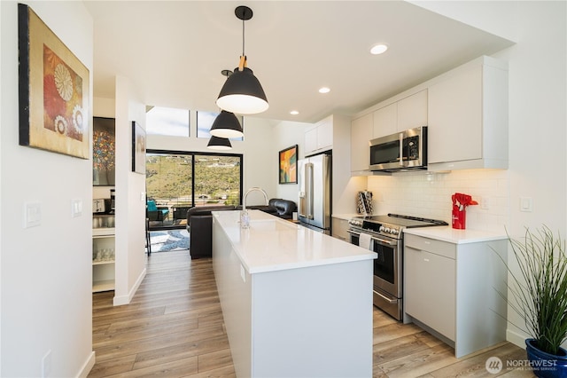 kitchen with stainless steel appliances, tasteful backsplash, light countertops, light wood-style floors, and an island with sink