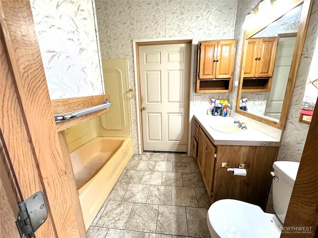 full bathroom featuring stone tile flooring, vanity, toilet, wallpapered walls, and a tub to relax in