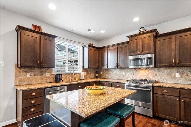 kitchen featuring dark wood finished floors, appliances with stainless steel finishes, dark brown cabinetry, a sink, and a kitchen breakfast bar