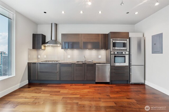 kitchen featuring wood finished floors, wall chimney range hood, appliances with stainless steel finishes, backsplash, and electric panel