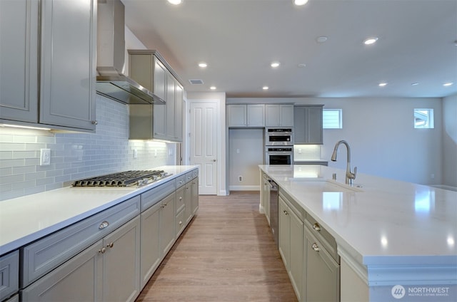 kitchen with light wood-style floors, a large island, stainless steel appliances, wall chimney range hood, and a sink