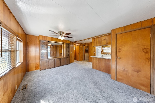 unfurnished living room with visible vents, wood walls, a ceiling fan, and light colored carpet