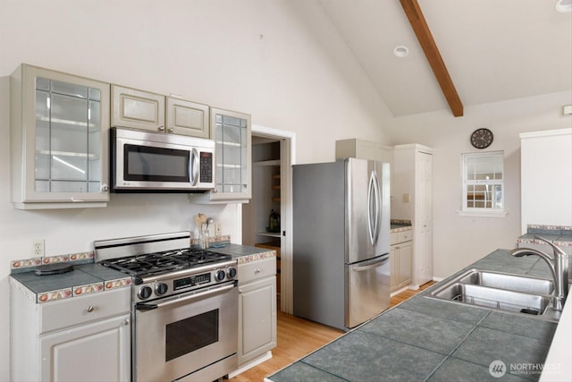 kitchen with beam ceiling, light wood-style flooring, appliances with stainless steel finishes, glass insert cabinets, and a sink