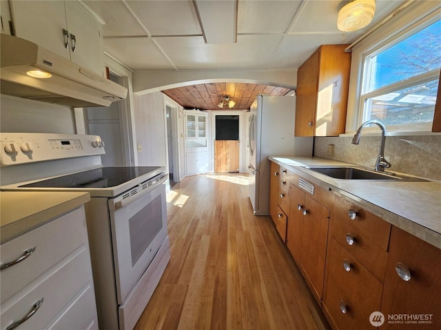 kitchen with under cabinet range hood, white appliances, a sink, light wood finished floors, and brown cabinetry