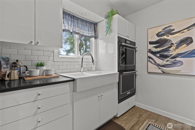 kitchen featuring wood finished floors, a sink, white cabinets, decorative backsplash, and multiple ovens