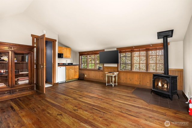 unfurnished living room featuring dark wood finished floors, wainscoting, a wood stove, vaulted ceiling, and wood walls