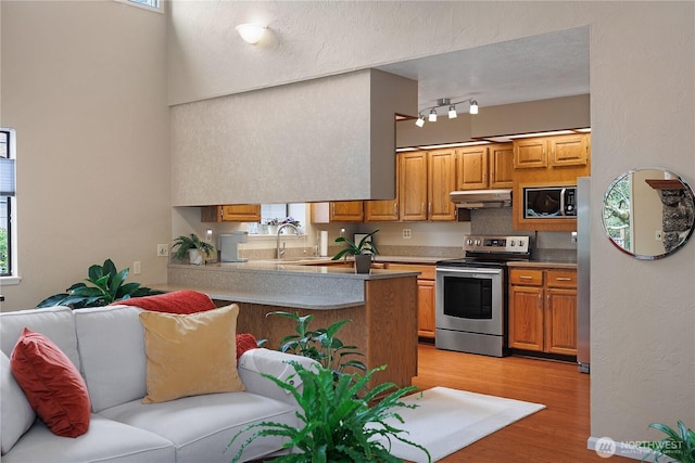 kitchen with under cabinet range hood, light wood-type flooring, a peninsula, brown cabinetry, and stainless steel appliances