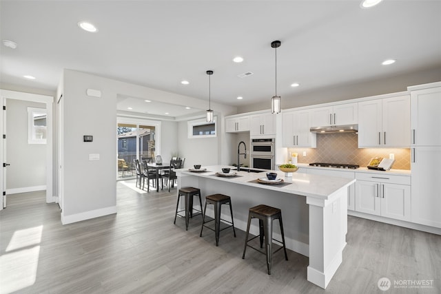 kitchen with backsplash, double oven, gas cooktop, an island with sink, and under cabinet range hood