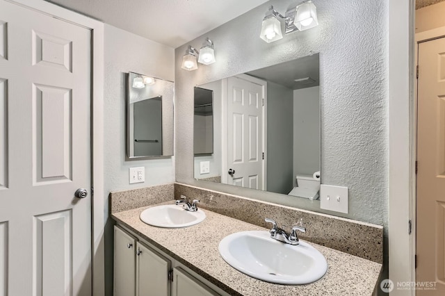 bathroom with a textured wall, a sink, toilet, and double vanity