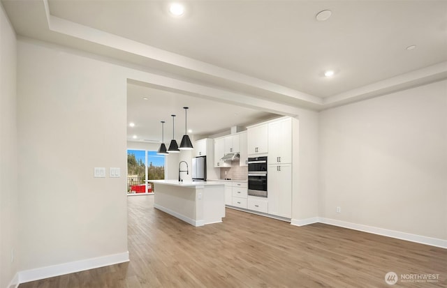 kitchen featuring light wood finished floors, a center island with sink, decorative backsplash, white cabinetry, and a sink