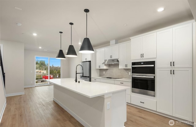 kitchen with double wall oven, freestanding refrigerator, an island with sink, under cabinet range hood, and black electric cooktop