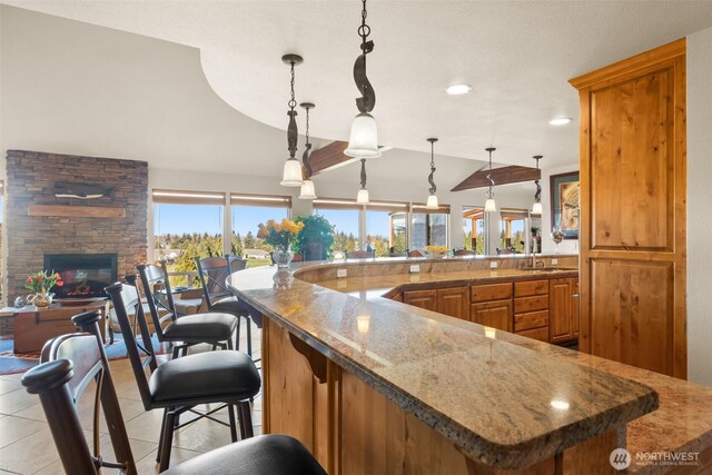 kitchen featuring light tile patterned floors, brown cabinets, vaulted ceiling, a stone fireplace, and pendant lighting