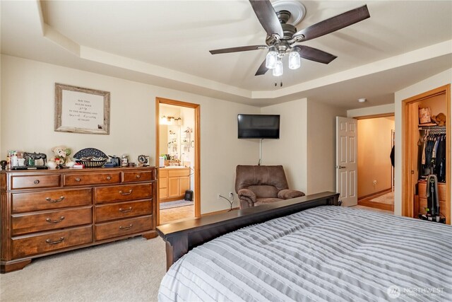 bedroom featuring a raised ceiling, ensuite bathroom, a ceiling fan, baseboards, and light colored carpet