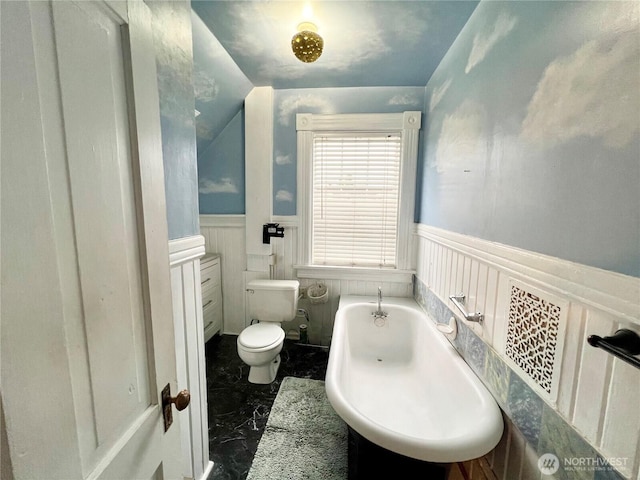 bathroom featuring marble finish floor, toilet, wainscoting, vaulted ceiling, and a tub