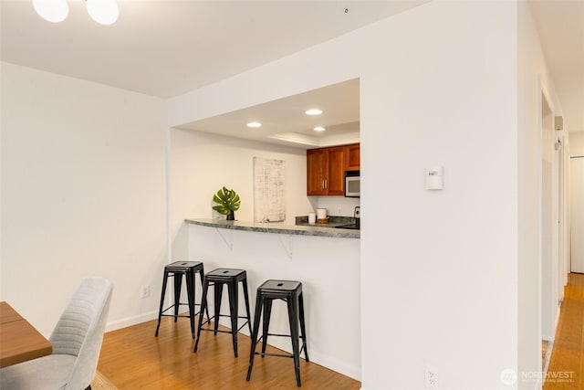 kitchen with recessed lighting, light wood-type flooring, brown cabinetry, stainless steel microwave, and a kitchen bar