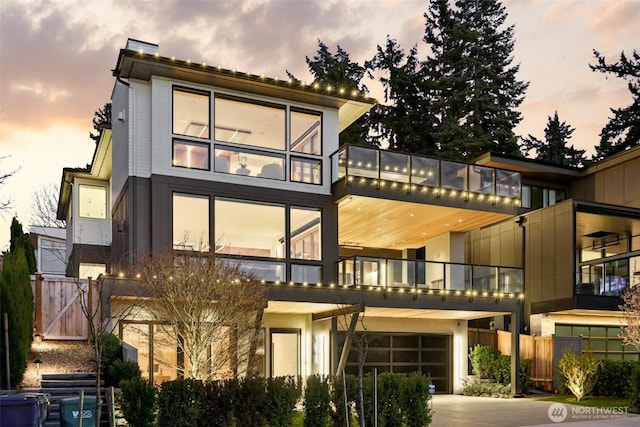 back of house at dusk featuring driveway, a chimney, stucco siding, fence, and a garage