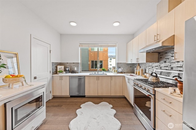 kitchen with under cabinet range hood, light brown cabinetry, light countertops, appliances with stainless steel finishes, and a sink