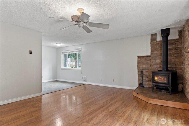 unfurnished living room with a ceiling fan, a wood stove, a textured ceiling, and wood finished floors