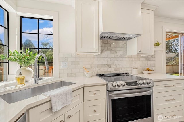 kitchen with a sink, electric stove, wall chimney range hood, light countertops, and decorative backsplash