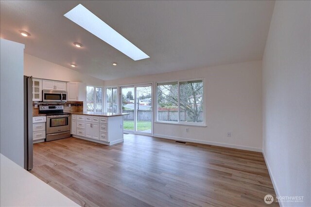 kitchen with light wood finished floors, lofted ceiling with skylight, appliances with stainless steel finishes, a peninsula, and white cabinets