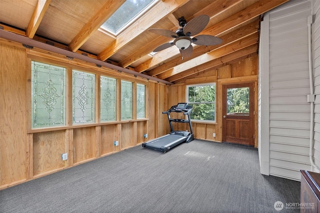 exercise room featuring carpet floors, vaulted ceiling with skylight, wooden walls, and a ceiling fan