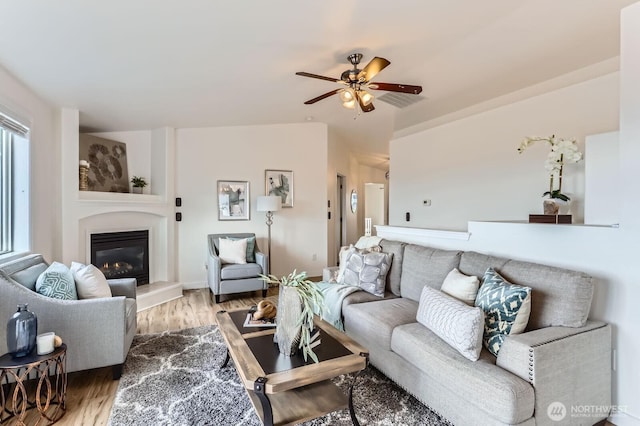 living room featuring light wood finished floors, visible vents, ceiling fan, vaulted ceiling, and a glass covered fireplace