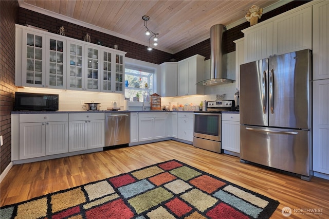 kitchen featuring stainless steel appliances, dark countertops, wall chimney range hood, brick wall, and wooden ceiling