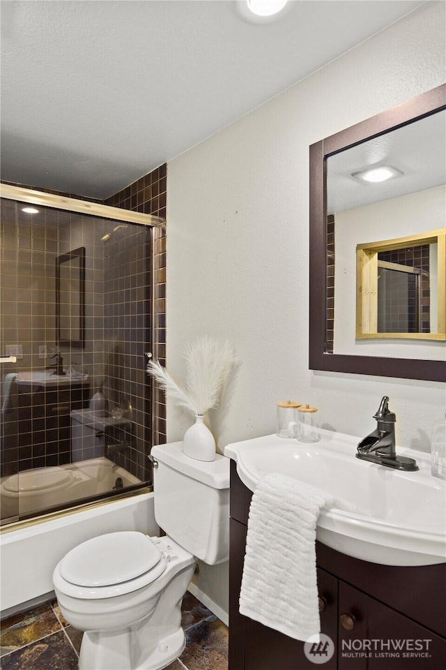 bathroom with toilet, vanity, baseboards, combined bath / shower with glass door, and stone tile flooring