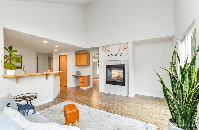 living room with baseboards, a high ceiling, recessed lighting, a tiled fireplace, and light wood-type flooring