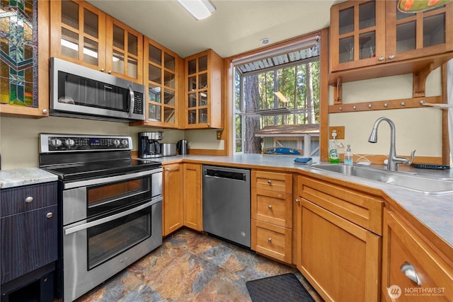 kitchen featuring a sink, light countertops, appliances with stainless steel finishes, stone finish flooring, and glass insert cabinets