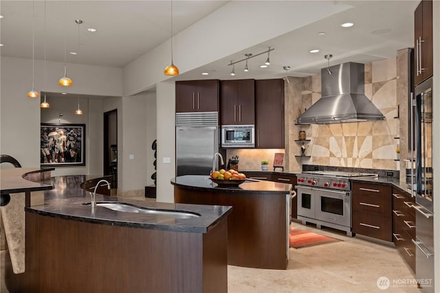 kitchen featuring decorative backsplash, built in appliances, a kitchen island with sink, wall chimney range hood, and a sink