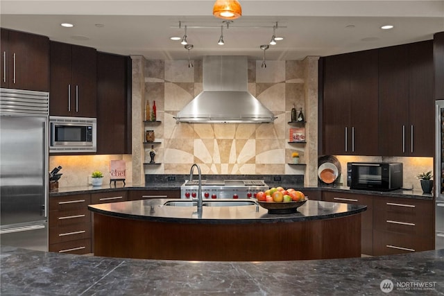 kitchen featuring built in appliances, wall chimney exhaust hood, dark stone counters, and decorative backsplash