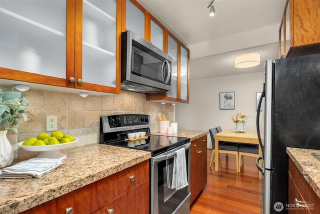 kitchen with stainless steel appliances, decorative backsplash, brown cabinetry, glass insert cabinets, and wood finished floors