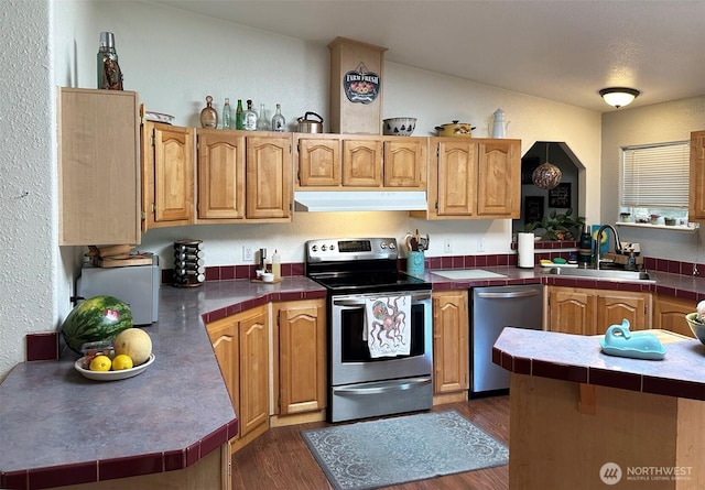 kitchen with tile countertops, under cabinet range hood, a sink, appliances with stainless steel finishes, and dark wood-style floors