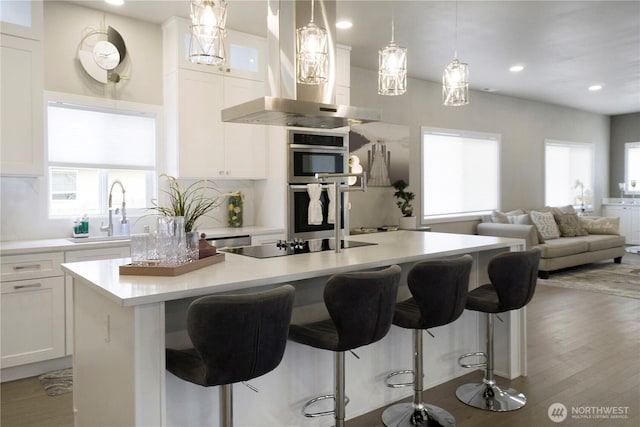 kitchen featuring an island with sink, wall chimney exhaust hood, open floor plan, black electric cooktop, and a sink