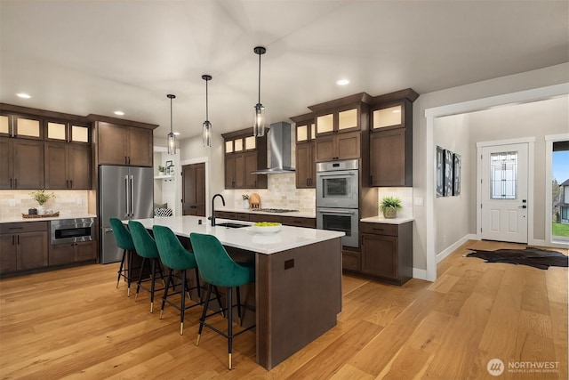 kitchen featuring light countertops, appliances with stainless steel finishes, a sink, wall chimney range hood, and dark brown cabinets