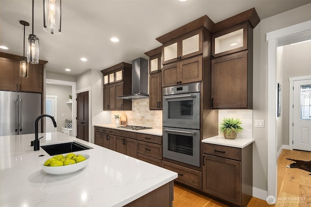 kitchen featuring decorative backsplash, appliances with stainless steel finishes, light wood-style floors, a sink, and wall chimney exhaust hood