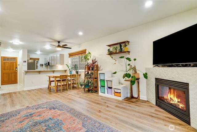 living area with a tiled fireplace, light wood-style floors, baseboards, and a ceiling fan