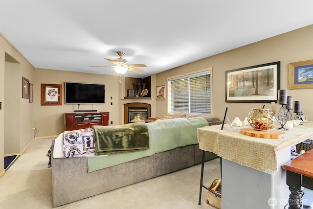 living area with a ceiling fan, a glass covered fireplace, light carpet, and baseboards