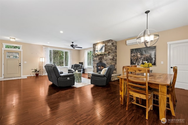 dining space featuring baseboards, a wall unit AC, wood finished floors, a stone fireplace, and ceiling fan with notable chandelier